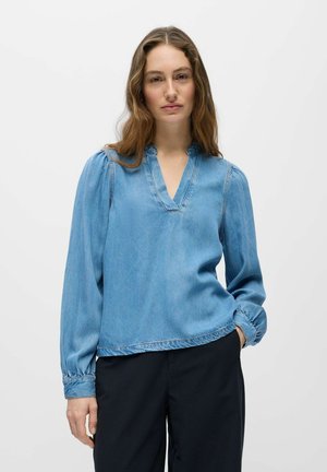 Femme aux longs cheveux bruns portant un chemisier en denim bleu clair à col en V avec des manches longues bouffantes et un pantalon noir, se tenant devant un fond blanc uni.
