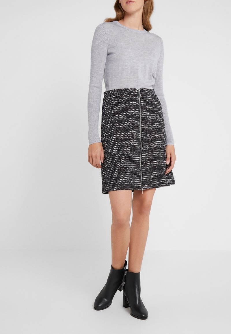 Femme portant un haut gris à manches longues, une jupe trapèze texturée noire et blanche avec fermeture éclair devant, et des bottines noires à talons blocs.