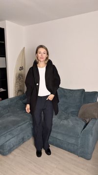 Casaco preto oversized com capuz e detalhe de botões, combinado com uma blusa branca e calças largas pretas; de pé num sofá azul-pavão.