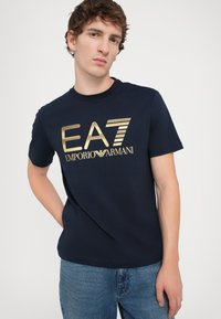 Tamsiai mėlynas medvilninis marškinėliai su auksiniu EA7 Emporio Armani logotipu ir horizontaliomis juostelėmis. Trumpomis rankovėmis ir apvalia iškirpte.