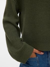 Groene gebreide trui met een textuurpatroon, geribde mouwen en een cropped design, gecombineerd met blauwe denim jeans.