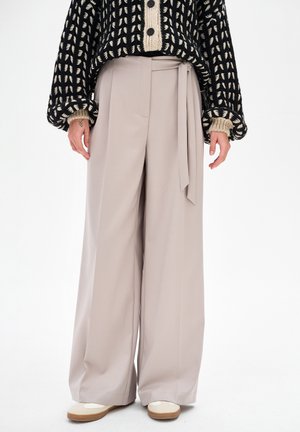 Beżowe spodnie z szerokimi nogawkami z wysokim stanem i paskiem w talii, zestawione z czarno-beżowym wzorzystym dzianinowym swetrem oraz białymi butami.