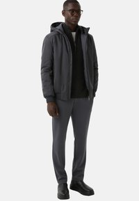 Mężczyzna w ciemnoszarej kurtce z kapturem, czarnej bluzie z zamkiem, szarych spodniach, czarnych butach i okularach, stojący na tle jasnego, jednolitego tła.