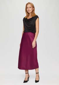 Zwarte coltrui gecombineerd met een vloeiende, knielange bordeaux satijnen rok. Model draagt zwarte hoge hakken met enkelbandjes.