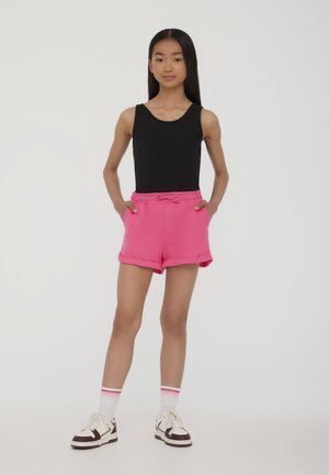 Canotta nera senza maniche, pantaloni corti in cotone rosa con elastico in vita e cordino, orli arrotolati, abbinati a calzini bianchi e scarpe da ginnastica.