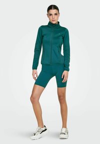 Femme debout, portant une veste zippée bleu sarcelle et un short de vélo assorti, avec des baskets blanches et noires, sur un fond uni.