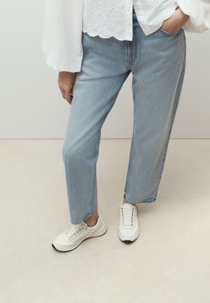 Person iført lyseblå jeans, hvid broderet bluse og hvide sneakers, der står på en lys gulv med den ene hånd i lommen.
