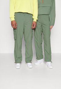 Olive kalhoty typu cargo s bočními kapsami, doplněné bílými teniskami. Jeden má na sobě žlutou mikinu s kapucí, druhý zelený proužkovaný top.