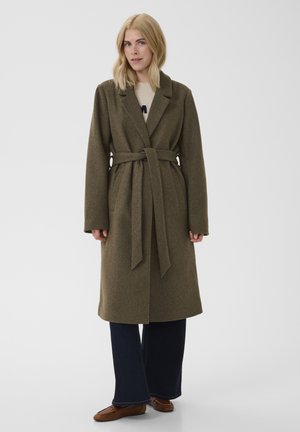 Femme portant un manteau en laine vert olive ceinturé, un pantalon large foncé, des ballerines marron, debout devant un fond clair uni.