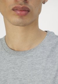 Grå T-skjorte med kort erm og rund hals, myk tekstur og synlige sømmer, iført av en person som kun viser hals og lepper.