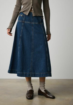 Mittellanger dunkelblauer Jeansrock mit vorderen Nähten, getragen mit grauen gerippten Socken, braunen Lederloafern und einem kurzen, gerippten, knopfbesetzten Cardigan.