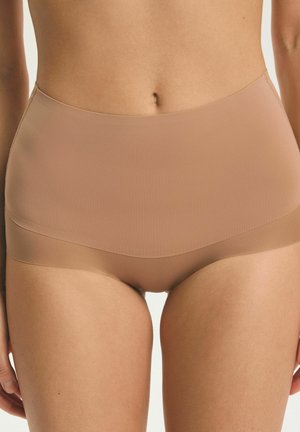 Žena nosi bezšvové telové nohavičky s vysokým pásom, ukazujúce brucho a hornú časť stehien na jednoduchom pozadí.