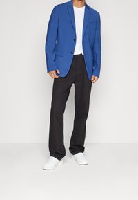 Blå skräddarsydd kavaj, vit t-shirt, svarta byxor och vita sneakers. Kavajen har slag och ett enda bröstfack.