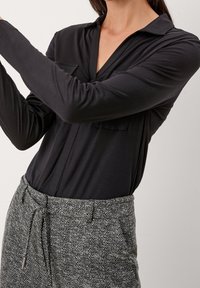 Camicia nera a maniche lunghe con colletto e due tasche frontali, abbinata a pantaloni grigi a spina di pesce con cordoncino.
