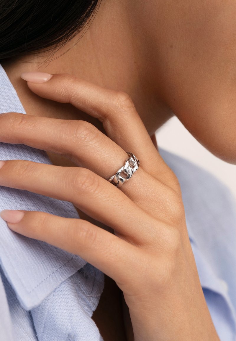 Silberner Ring mit einem verwobenen Kettendesign, vor einem hellblauen Stoffhintergrund. Glatte Textur mit polierter Oberfläche.