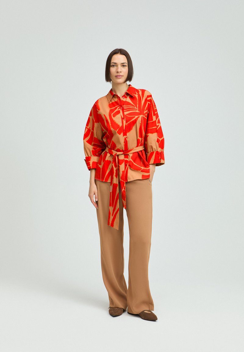 Rotes und beiges gemustertes Hemd mit einer Krawatte in der Taille, kombiniert mit beigen Hosen. Glattes Material, entspannte Passform und weitem Bein. Braune Schuhe.