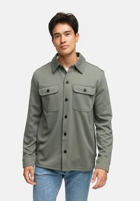 Grøn skjorte med knapper i struktureret stof, der har to brystlommer, en foldet krave og mørke knapper. Bæres med blå jeans.