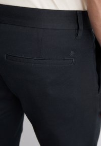 Pantaloni blu scuro con una texture liscia, dotati di una tasca posteriore e dettagli di cucitura sottili. Include un piccolo logo vicino alla tasca.