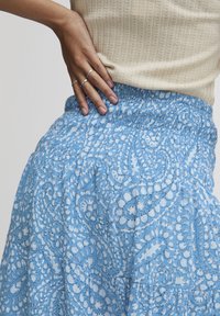 Fustă albastră deschisă cu un model complex de paisley alb, talie adunată și textură de material fluid. Top de nuanță pielii vizibil parțial deasupra.