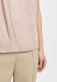 Frau in einem blassen rosa ärmellosen Oberteil und beigen hochgeschnittenen Hosen mit Plissee-Detail, stehend vor einem einfarbigen Hintergrund.