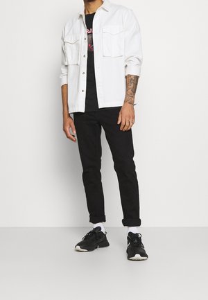 Witte denimjas met twee borstzakken over een zwart graphic t-shirt, gecombineerd met zwarte tapered jeans en zwarte sportschoenen.