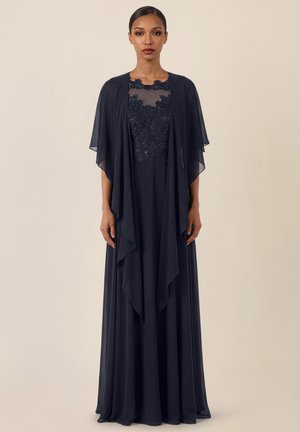 Frau trägt ein bodenlanges marineblaues Kleid mit Spitzenmieder und transparenten, drapierten Cape-Ärmeln, steht vor einem schlichten beigen Hintergrund.