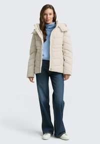 Chaqueta de plumón beige con cierre de cremallera y botones a presión, con un cuello alto. Combinada con un suéter de cuello alto azul claro y jeans de campana azules.