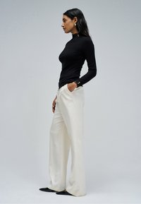Woman in black long-sleeve top, white wide-leg trousers, black pointed shoes, posing sideways with hands in pockets on neutral background.