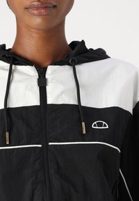 Cortavientos en blanco y negro con cremallera frontal, capucha y ribetes blancos; presenta un logotipo en el pecho y detalles metálicos en la cremallera.