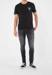 Czarna bawełniana koszulka z geometrycznym białym nadrukiem na piersi, w połączeniu z podniszczonymi szarymi dżinsami i czarnymi trampkami.