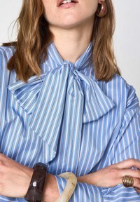 Blusa a righe blu e bianche con un grande fiocco al colletto. Tessuto morbido con una vestibilità larga. Braccialetti marroni e crema sul polso.