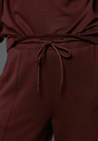 Set de pijamale bordo care include un top lejer și pantaloni cu talie elastică prevăzuți cu șnur de ajustare legat. Țesătură netedă și cusături minimaliste.
