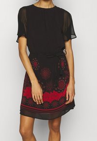 Černé šaty s krátkými průsvitnými rukávy, červenými květinovými vzory a pasem se stažením na šňůrku. Spodní část zdobená vrstveným červeným vzorem. Měkká, splývavá látka.