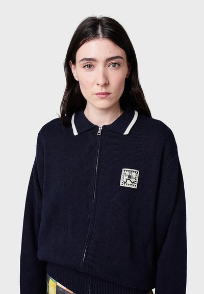 Marinblå zip-up tröja med ribbat mönster, vita krageaccenter och en fyrkantig logotypapplikation med ett mönster. Kortad passform med en strukturerad form.