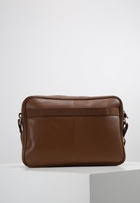 Brązowa skórzana prostokątna torba na ramię z poziomym paskiem z tyłu i regulowanymi klamrami po bokach, umieszczona na białej powierzchni.