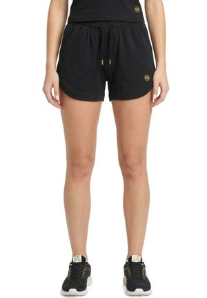 Modèle portant un short de sport noir à cordon avec des détails dorés et des baskets noires sur fond blanc.