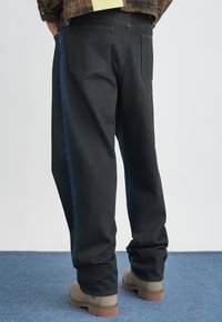 Črne jeans hlače z sproščenim krojem, z temno modrim stranskim panelom, petimi žepi in ravnim krojem. V kombinaciji z bež obutvijo.