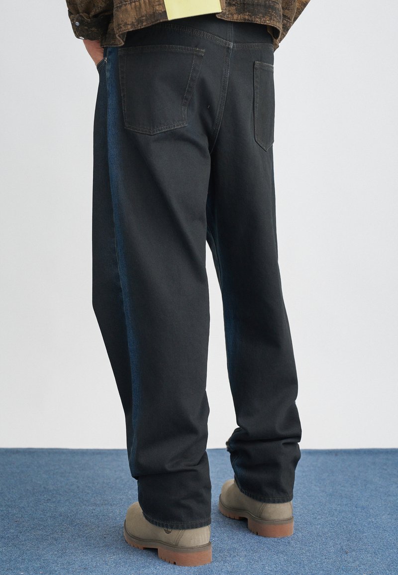 Črne jeans hlače z sproščenim krojem, z temno modrim stranskim panelom, petimi žepi in ravnim krojem. V kombinaciji z bež obutvijo.