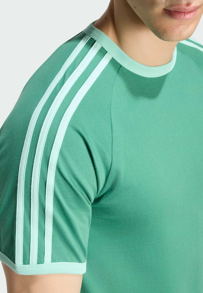 Chemise verte à manches courtes avec trois bandes vert clair allant de l’épaule au bord de la manche sur le torse supérieur d’une personne debout.