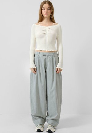 Junge Frau trägt ein weißes, geripptes, schulterfreies, kurzes Oberteil, hellgraue, weit geschnittene Nadelstreifenhose und weiße Turnschuhe, steht vor einem schlichten Hintergrund.