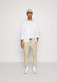Beige chinos with elastic cuffs, white t-shirt, layered white button-up shirt, and beige cap. White sneakers complete the outfit.