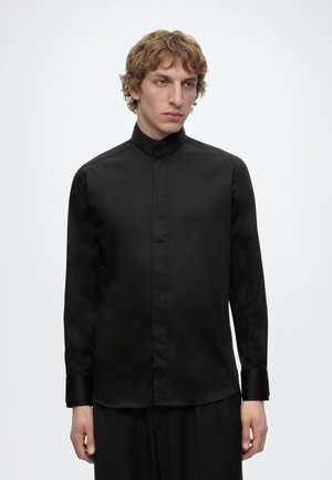 Ung mand med krøllet blondt hår iført en tætsiddende sort skjorte med høj krave og knapper samt sorte bukser, stående mod en ensartet hvid baggrund.