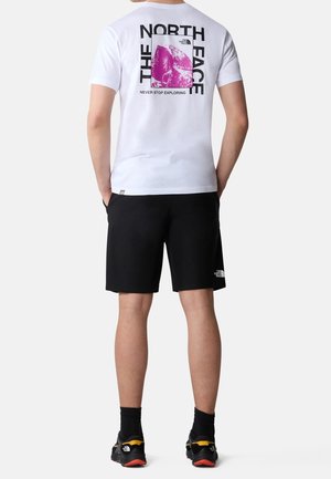 Bílý tričko s krátkým rukávem se vzorem růžové hory a nápisem na zádech. V kombinaci s černými šortkami a sportovními botami.