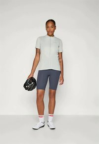 Ανοιχτό γκρι μπλούζα ποδηλασίας με κοντά μανίκια, μπροστινό φερμουάρ και υφή υφάσματος, συνδυασμένη με ναυτικές σορτς. Κρατάει ένα μαύρο κράνος, άσπρα παπούτσια και κάλτσες με κόκκινες λεπτομέρειες.