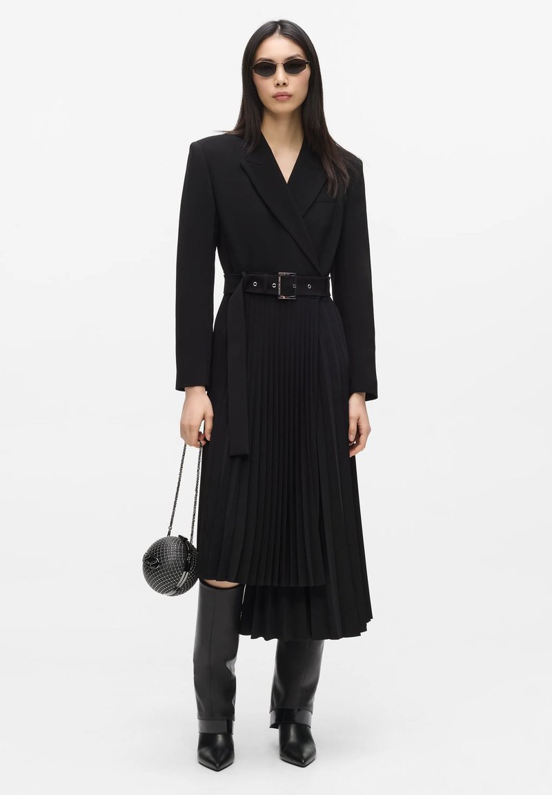 Γυναίκα που φοράει μαύρο blazer φόρεμα με ζώνη και πλισέ φούστα, μαύρες μπότες μέχρι το γόνατο, οβάλ γυαλιά ηλίου και κρατά μια μικρή στρογγυλή μαύρη τσάντα.