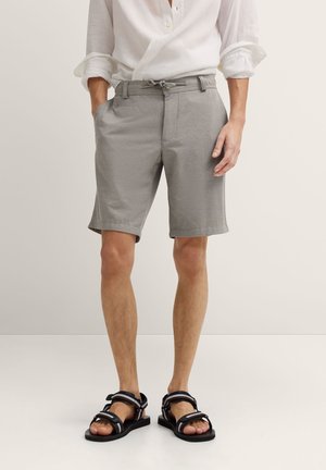 Mann trägt hochgekrempeltes weißes Hemd, graue knielange Shorts mit Kordelzug und schwarze Sandalen mit Riemen, steht mit teilweise in die Taschen gesteckten Händen.