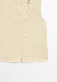 Beige stickad ärmlös topp med en rad tygklädda knappar som löper vertikalt nära sidsömmen.