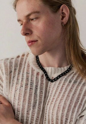 Person mit langen blonden Haaren, die einen transparenten weißen Strickpullover, eine schwarze Kettenkette und mehrere Ohrringe trägt und nach unten zur Seite schaut.
