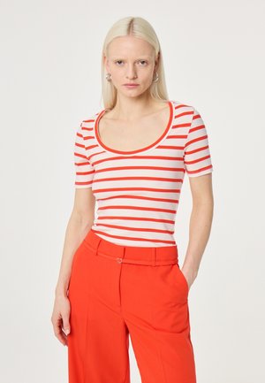 Woman with straight blonde hair wears a white shirt with red horizontal stripes and red high-waisted pants, standing with one hand in pocket.