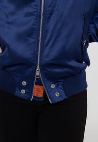 Marineblaue Bomberjacke mit glänzender Oberfläche, silbernem Reißverschluss, gerippten Bündchen und Saum; besitzt ein sichtbares oranges Etikett im Inneren.